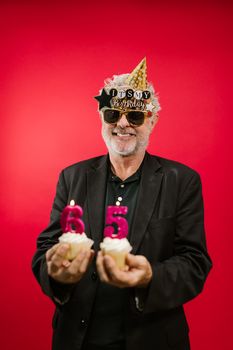 Cheerful senior man in a black suit holding cupcakes with '65' candles, celebrating his birthday.