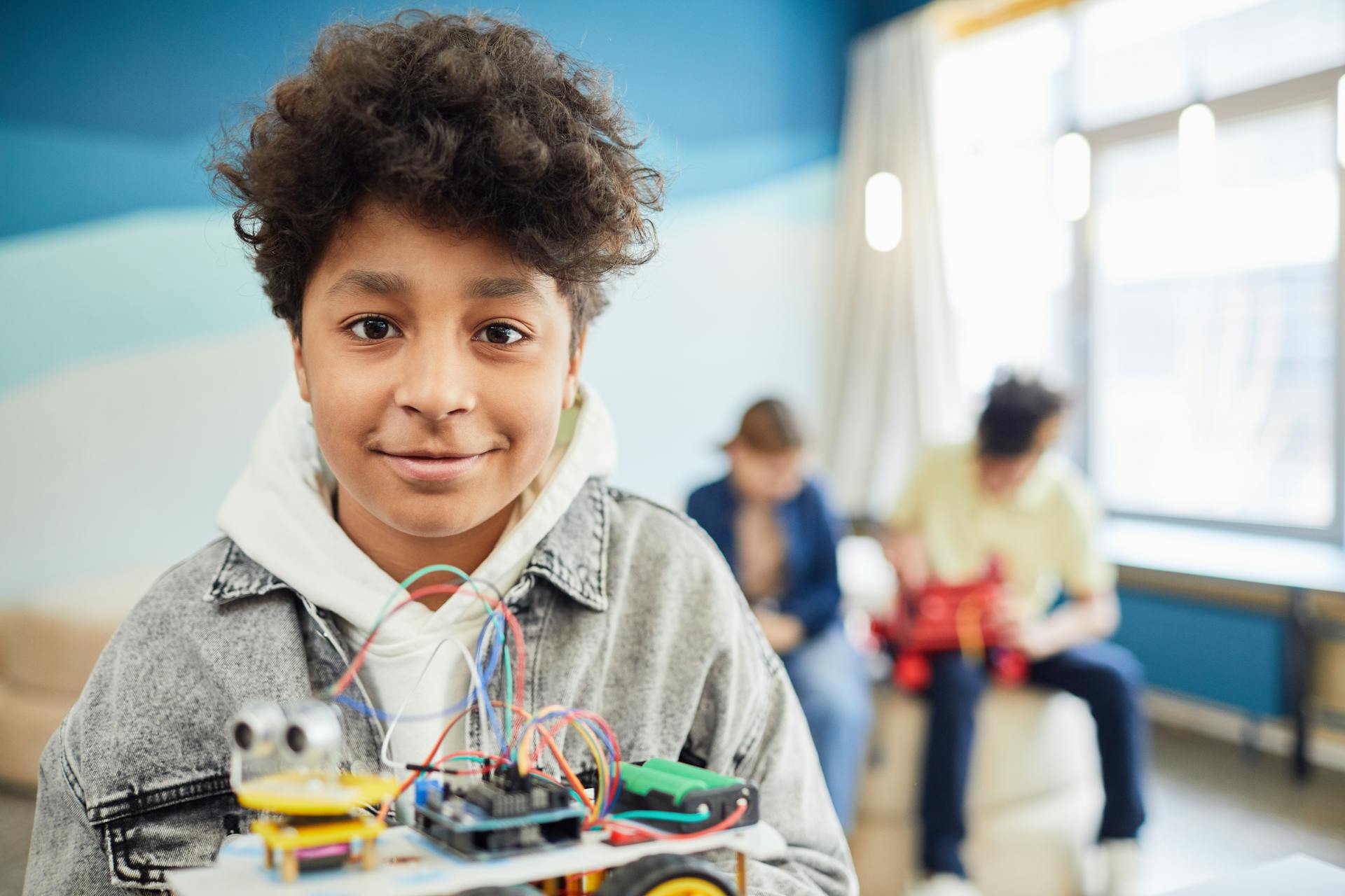 A young boy smiling confidently while holding a DIY robotics project in a classroom setting.