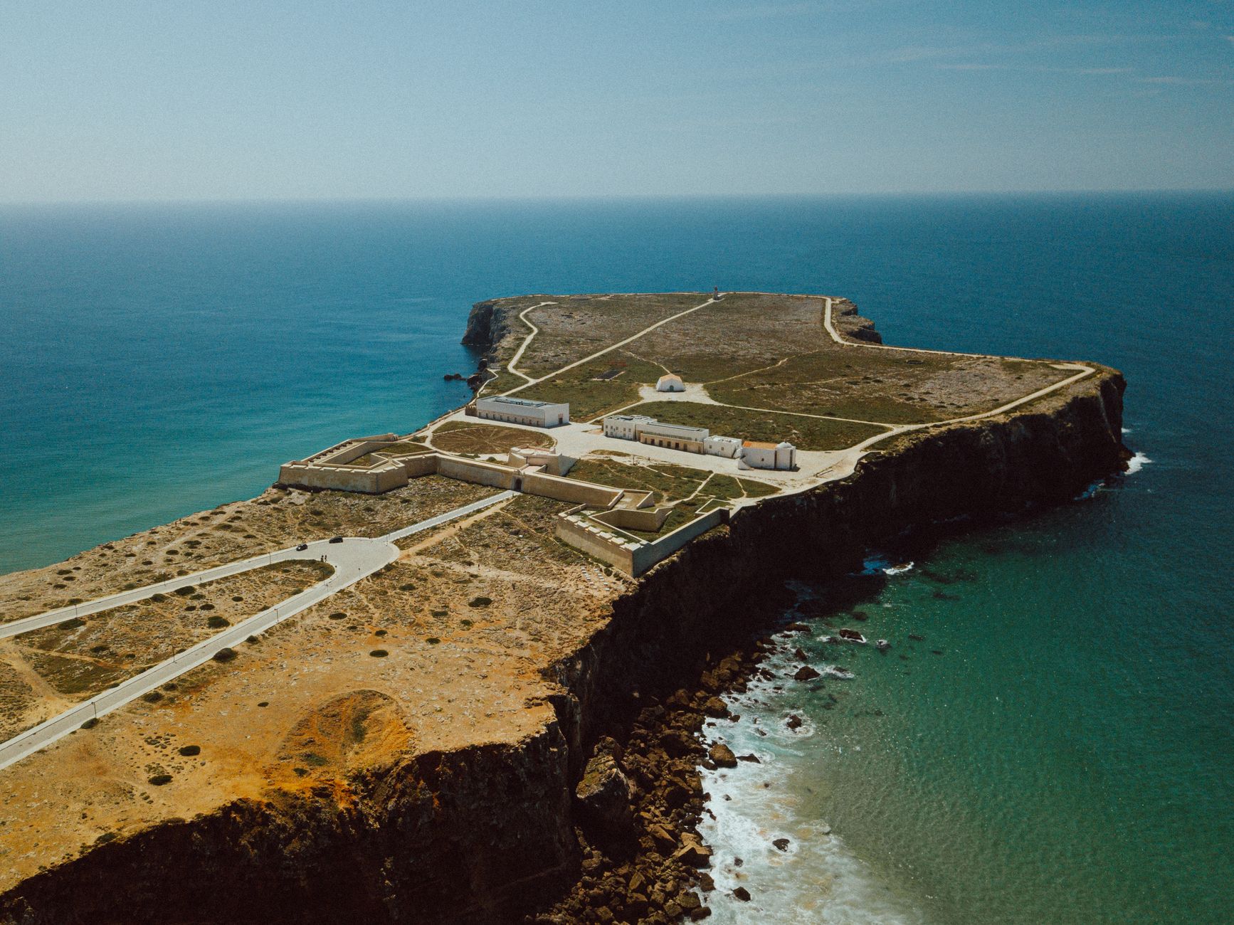 An aerial view of the Sagres promontory with the low walls and white buildings of the Fortaleza de Sagres atop sheer Atlantic cliffs