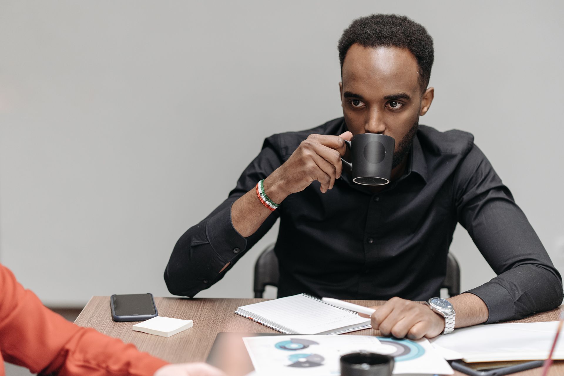 Man in a black shirt drinking coffee while sitting at an office table with documents nearby.