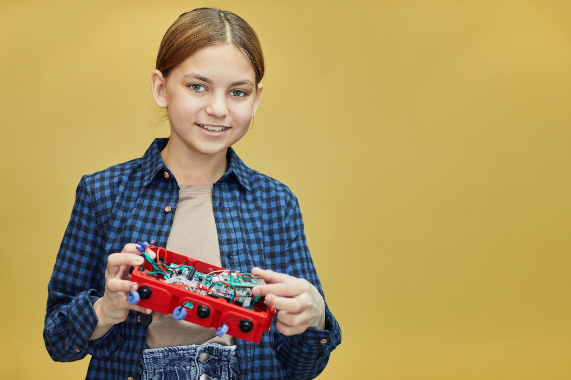 Caucasian girl happily holds a robotics kit against a yellow backdrop.