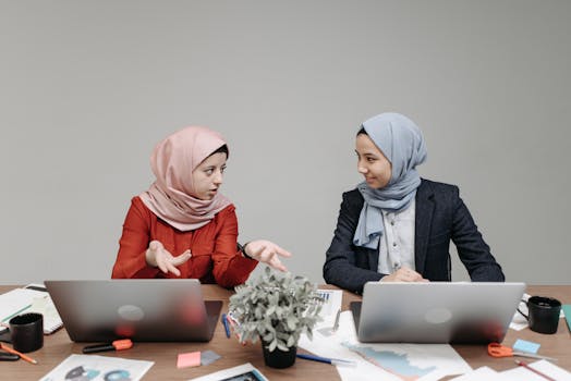 Two women in hijabs discussing business at an office table with laptops.