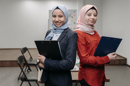 Two women in business attire with hijabs holding clipboards, symbolizing teamwork and professionalism.