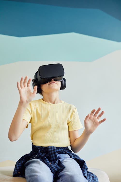Excited little girl in casual outfit wearing VR headset and sitting on sofa in light room
