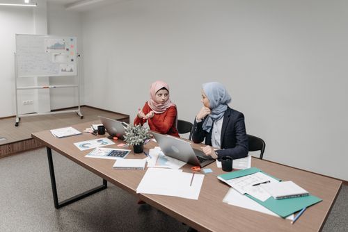 Free Two women in hijabs collaborating in a modern office setting with laptops and documents. Stock Photo