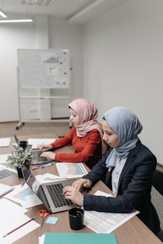 Two women in hijabs working on laptops in a contemporary office setting.