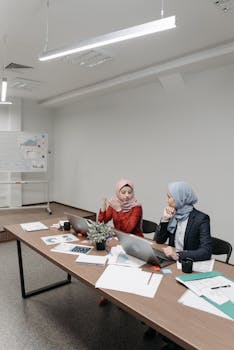 Two women in hijabs working on laptops in a modern office setting, discussing ideas.