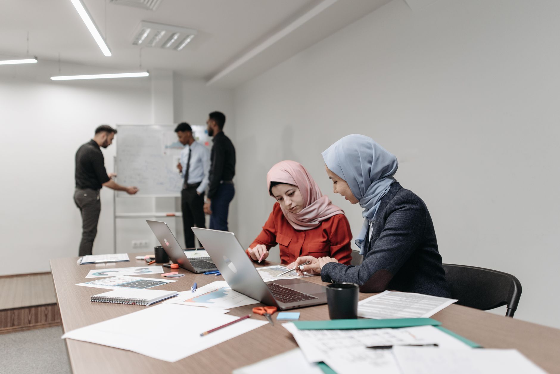 Young Muslim women in hijabs working at laptops in a training room, with colleagues collaborating at a whiteboard behind them
