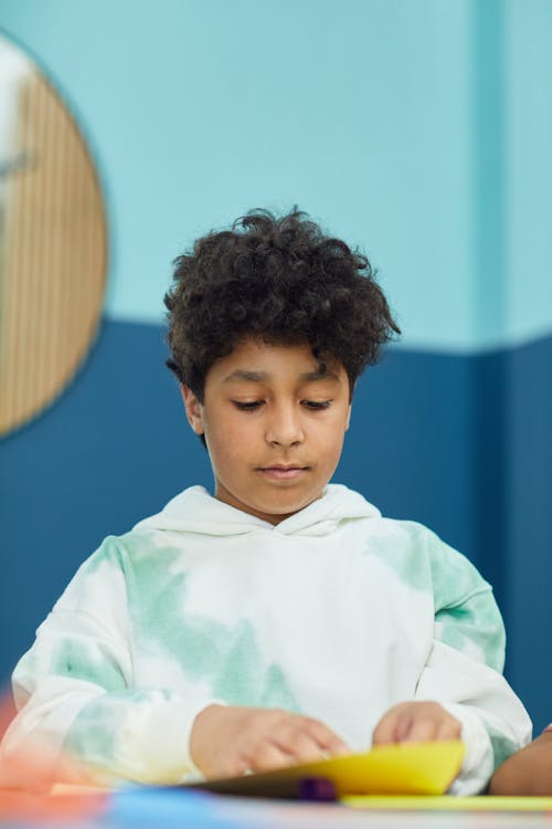 Free Concentrated ethnic schoolchild with curly hair in hoodie sitting at table and creating artwork in light room Stock Photo
