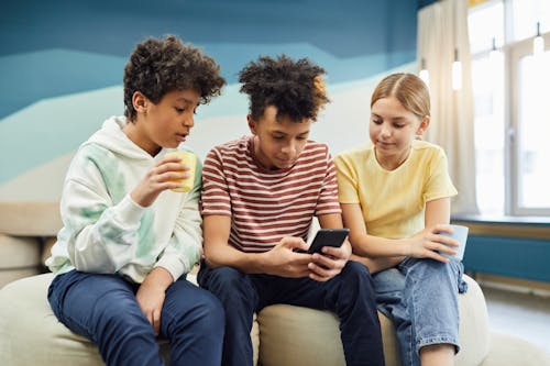 Free Group of multiracial children using smartphone while sitting on soft armchairs and holding cup of beverage in living room Stock Photo