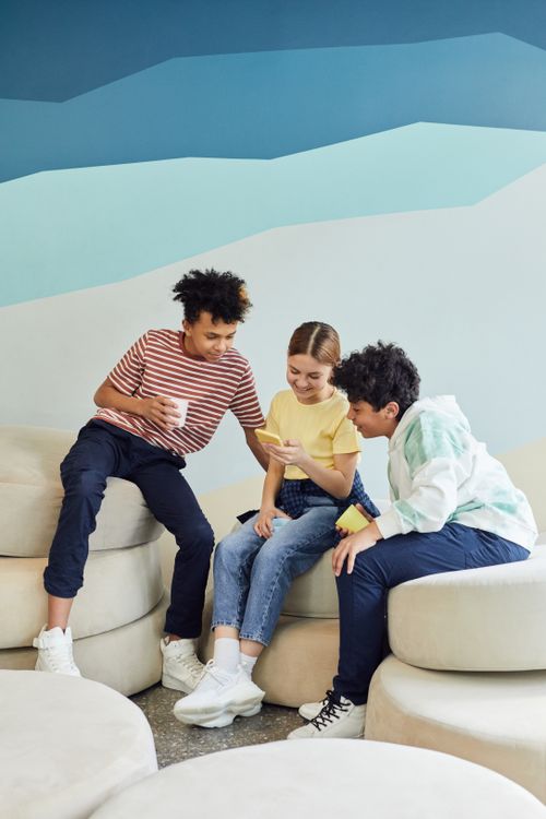 Free Group of diverse happy children sitting on cozy armchairs and using smartphone while holding cups of drink and looking at screen in light room Stock Photo