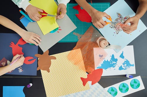 Free Top view of group of crop anonymous children sitting at table and creating colorful draft and artworks with paper and scissors Stock Photo