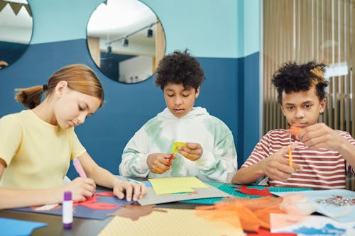 Free Group of concentrated multiracial children making artworks with colorful paper and scissors while sitting at table in light room Stock Photo