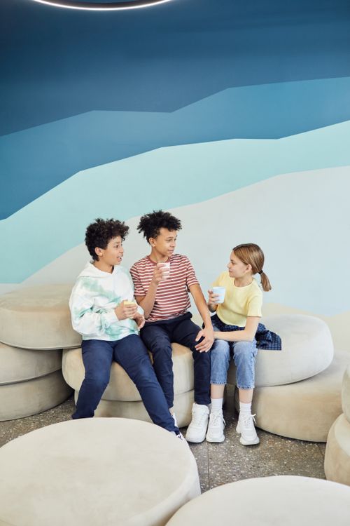 Free Group of excited multiethnic children sitting on chairs and talking with each other while holding cups of beverage in light room Stock Photo