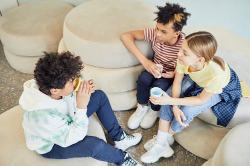 Free From above of group of multiethnic children sitting on soft pillows and drinking hot beverage while spending time together in living room Stock Photo