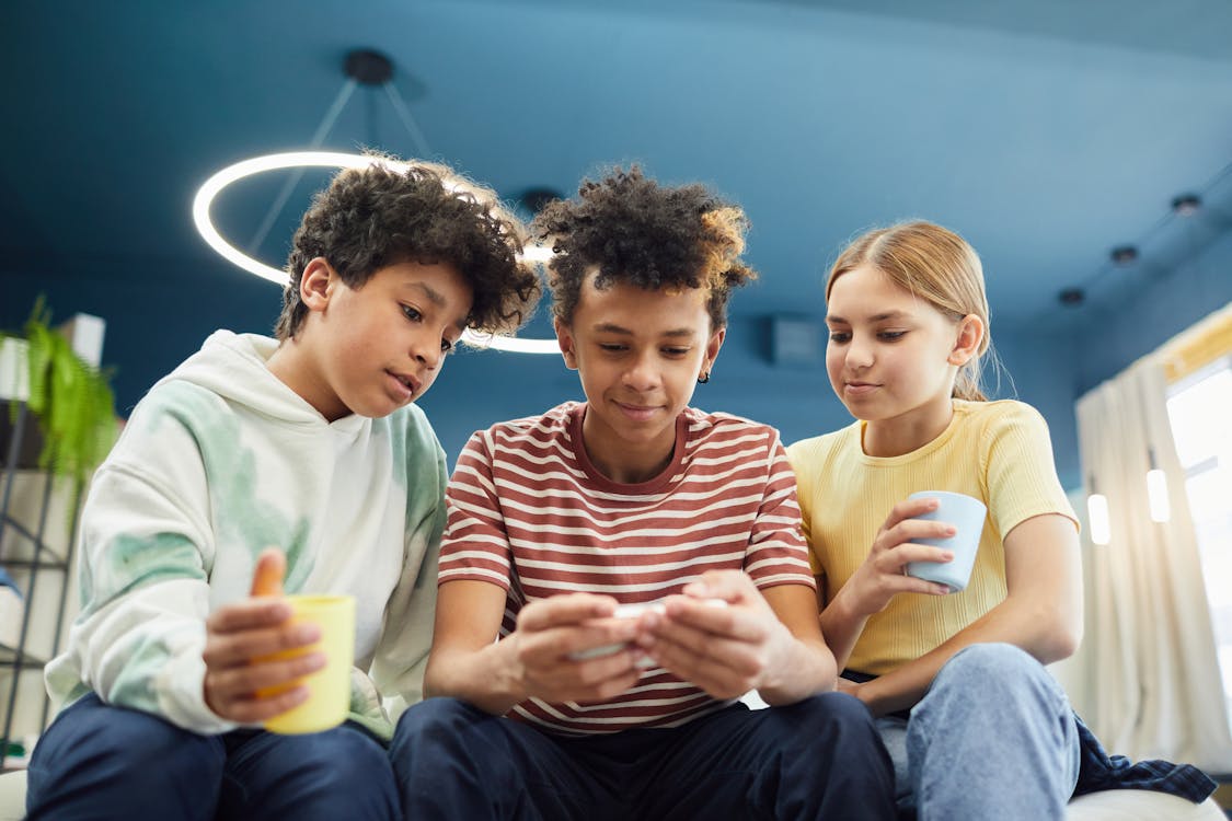 From below of group of diverse children spending time together while using smartphone and drinking beverages in living room