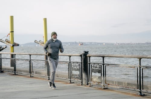 Full body of concentrated bearded African American male athlete wearing warm activewear running on quay near sea in sunny morning
