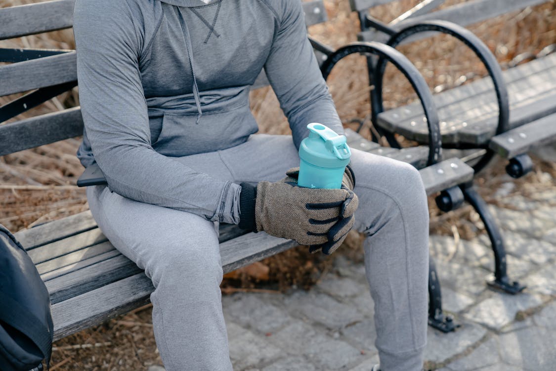 Crop unrecognizable fit male in gray warm sportswear and gloves sitting on wooden bench in park and holding bottle of water in hands in daylight