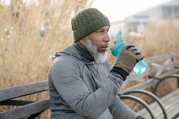 Old Black Man Sitting On Bench In Park And Drinking Water