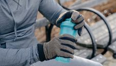 Crop anonymous male in gray activewear and gloves sitting on bench in park and opening bottle of water while resting after training in daytime