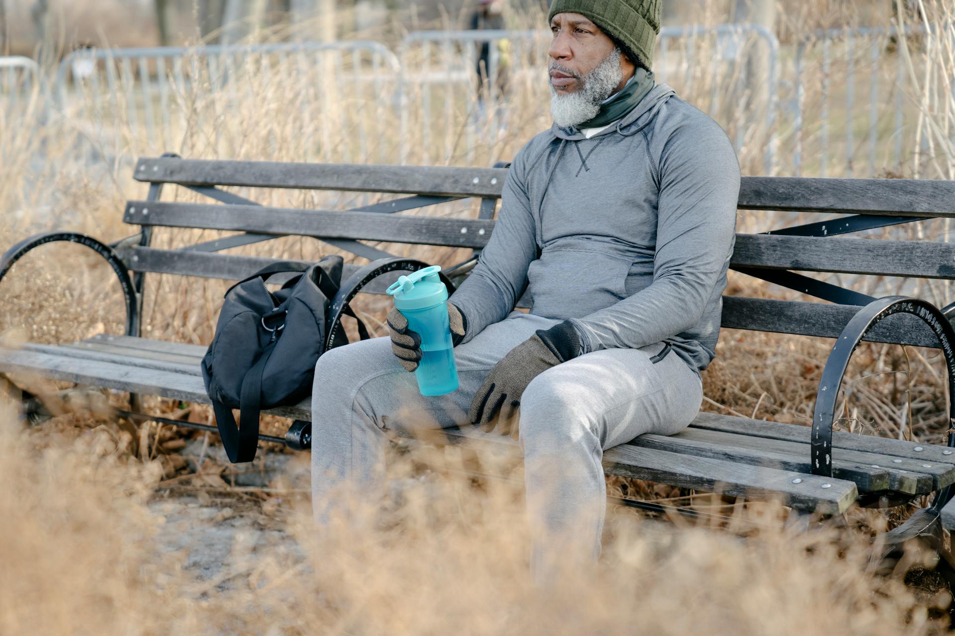 Side view of senior African American male wearing warm sportswear sitting on wooden bench with sport bag and bottle of water in city park and looking away pensively in daylight