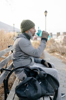 A senior man enjoys a drink while resting on a park bench during outdoor exercise.
