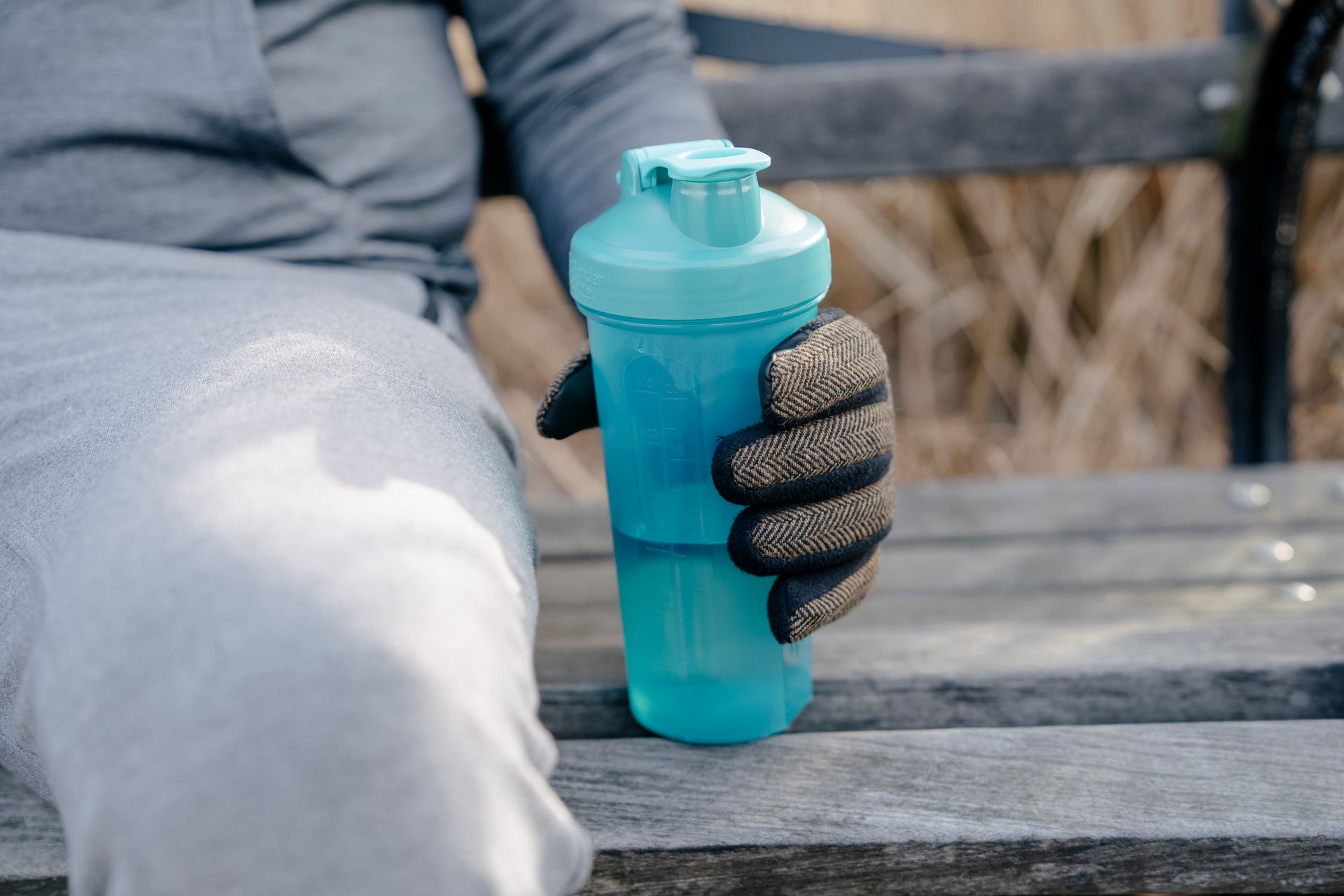 Crop anonymous male in activewear and gloves sitting on wooden bench in city park and holding bottle of water in hand in daylight