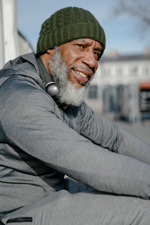 Free Confident African American senior beard male wearing gray sports clothes and knitted hat and headphones on neck sitting on street in sunny day Stock Photo