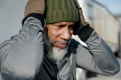 Bearded African American male wearing warm gray hoodie brown knitted gloves and green hat touching head while fixing headphones