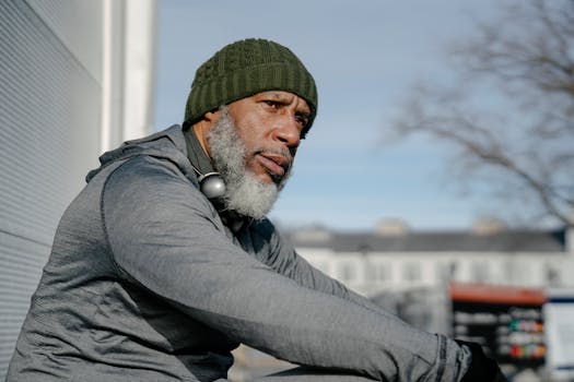 Senior African American man in a hoodie and beanie, sitting outdoors on a sunny day.