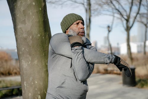 Serious bearded African American male in warm activewear stretching side muscle of arm near tree in park while doing workout