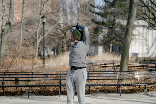 Concentrated African American male in casual outerwear standing in park and stretching triceps of hand while doing workout