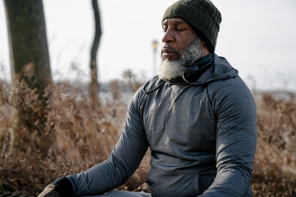 Free Bearded African American male in warm activewear and knitted hat sitting and meditating with closed eyes in autumn nature Stock Photo