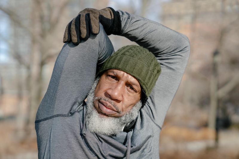 A senior African American man in a cap and gloves stretches in a park during fall, staying active for his lung and heart health.