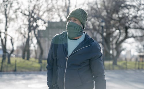 Male in warm clothes standing on street with face covered by scarf on blurred background of leafless trees