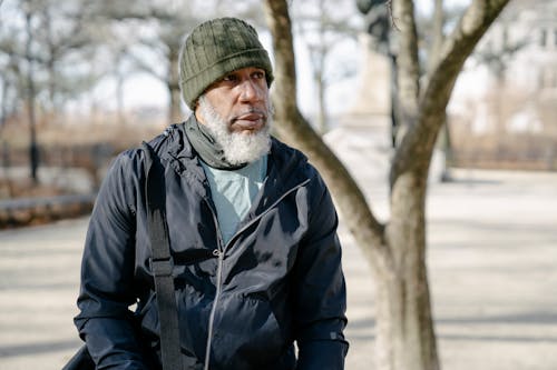 African American aged bearded male in warm jacket and cap looking away during stroll in autumn day