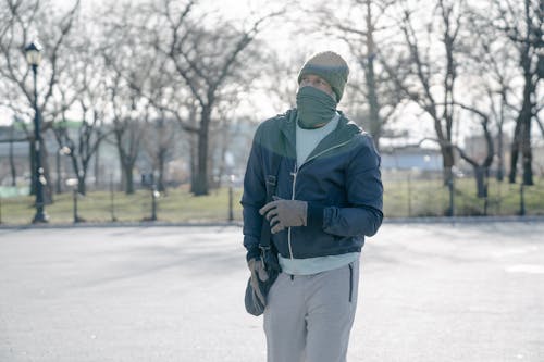 Unrecognizable male covering face with scarf on street with leafless trees and looking away