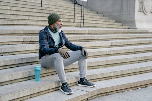 Full body of African American mature male wearing knitted hat and activewear and sneakers sitting on stairs with fitness bottle and looking away