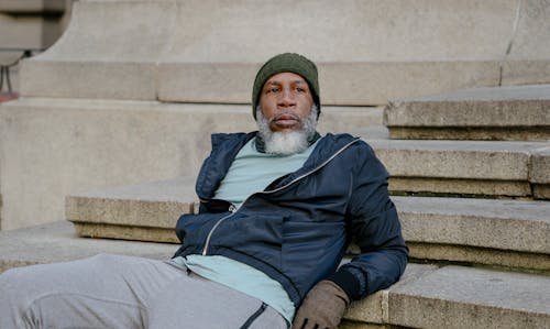 Senior bearded African American male in gray sweatpants and sport windbreaker jacket and knitted hat and gloves reclining on stone stairs