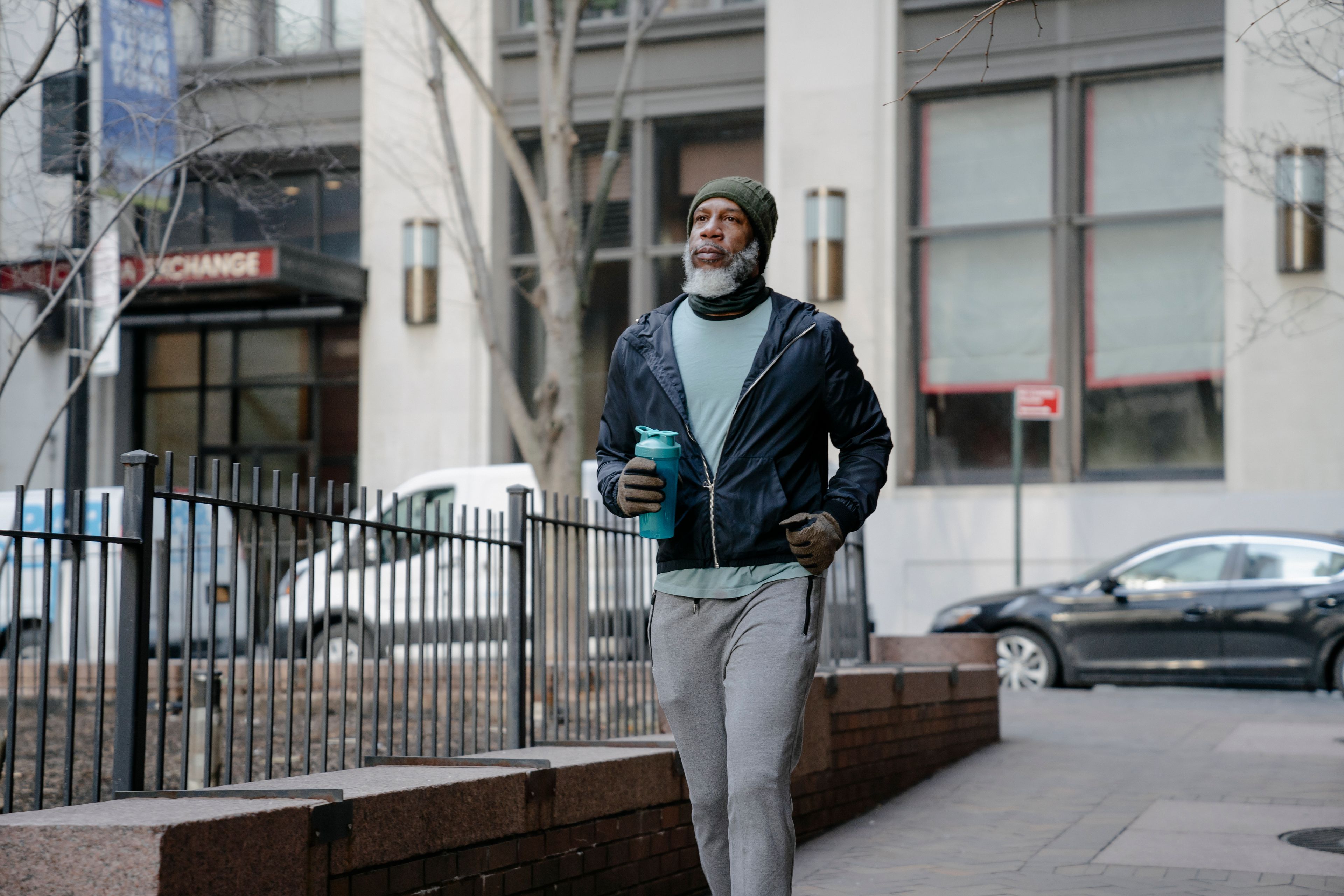 Concentrated elderly bearded black man in sportswear with fitness bottle jogging along metal fence in center of city near office buildings