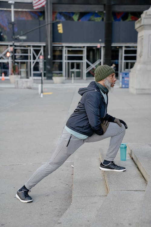 Side view of full length of mature beard African American male in windbreaker sport jacket and sweatpants and sneakers with fitness bottle stretching legs on step of stairs