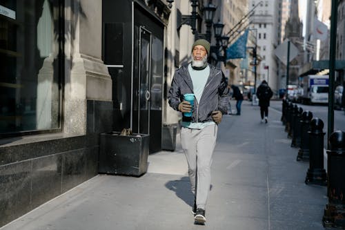 Free Full body of confident bearded African American male in activewear and warm gloves and hat walking along sidewalk with fitness bottle for water in sunny morning Stock Photo