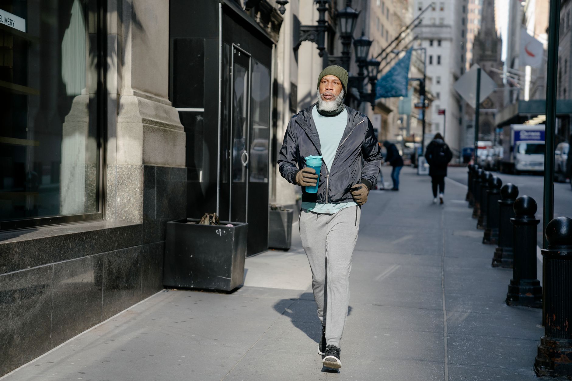 Full body of confident bearded African American male in activewear and warm gloves and hat walking along sidewalk with fitness bottle for water in sunny morning