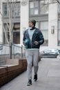 Full body of mature African American male wearing windbreak jacket and sweatpants and sneakers and knitted hat jogging along fence in sidewalk of city with fitness bottle in hand