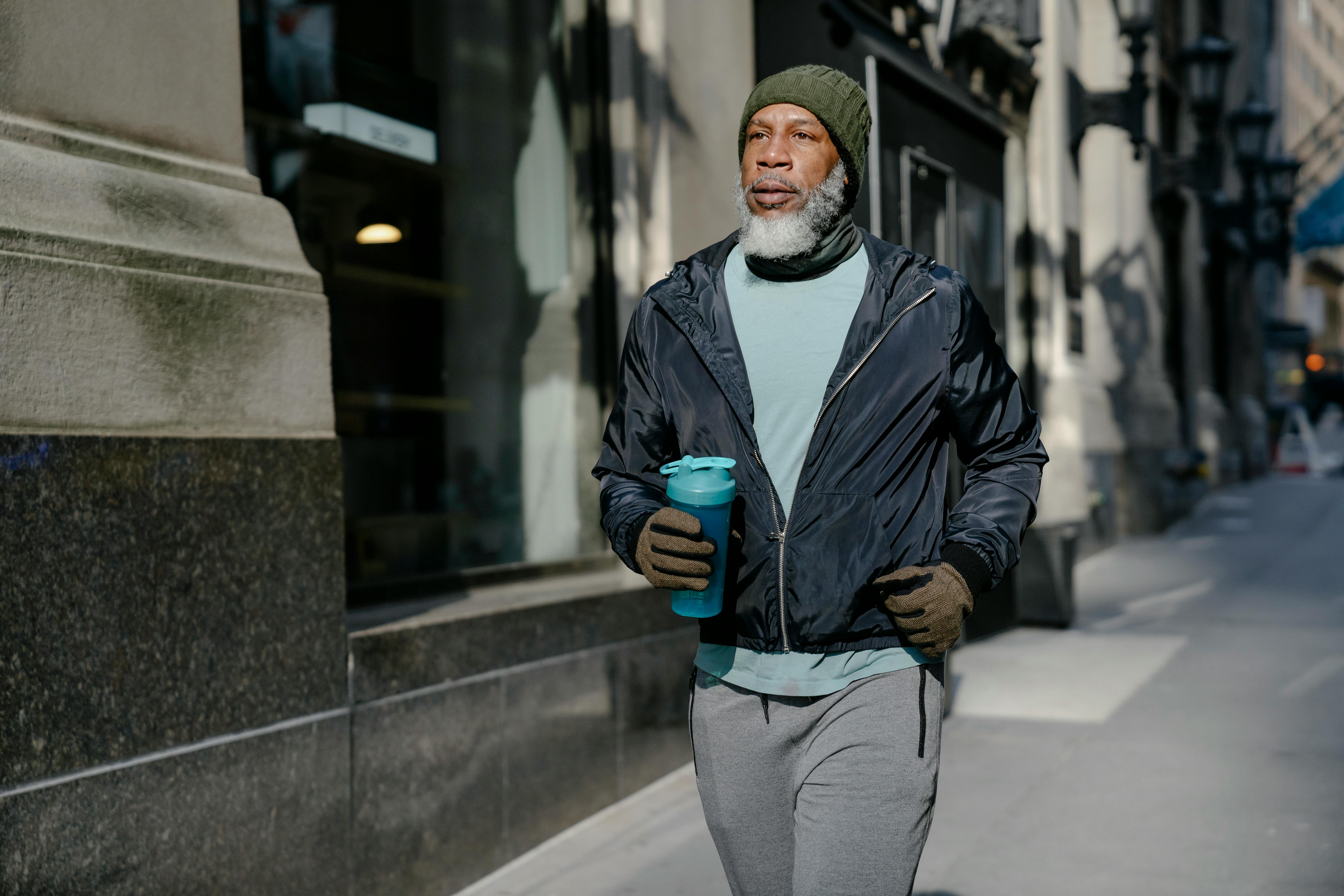 Free Serious elderly African American male in activewear and knitted hat and gloves running on pavement near building with sport bottle Stock Photo