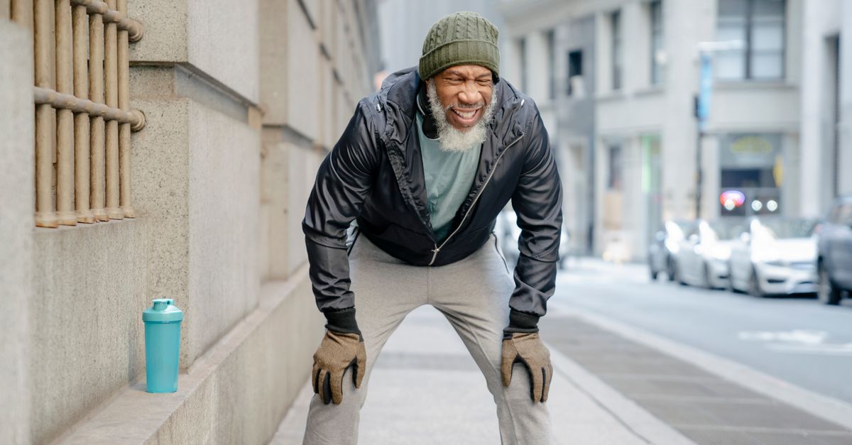 Concentrated bearded mature black man in activewear standing on street leaning forward with hands on knee while resting after hard training