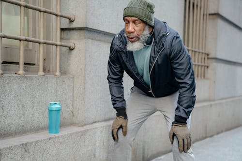 Concentrated bearded senior black man wearing warm sportswear leaning forward with hands on knees and closed eyes in street near building while break in workout