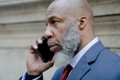 Concentrated bald bearded African American male entrepreneur wearing classy suit and speaking on smartphone in street in daytime