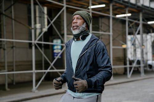 Serious African American male in warm hat and gloves with headphones on neck and windbreaker jacket running along metal protective construction in street