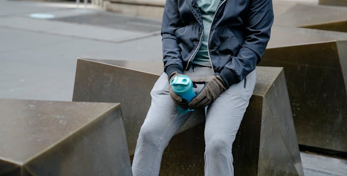 Crop unrecognizable male in jacket and sweatpants and gloves with fitness bottle sitting on stone parking barrier